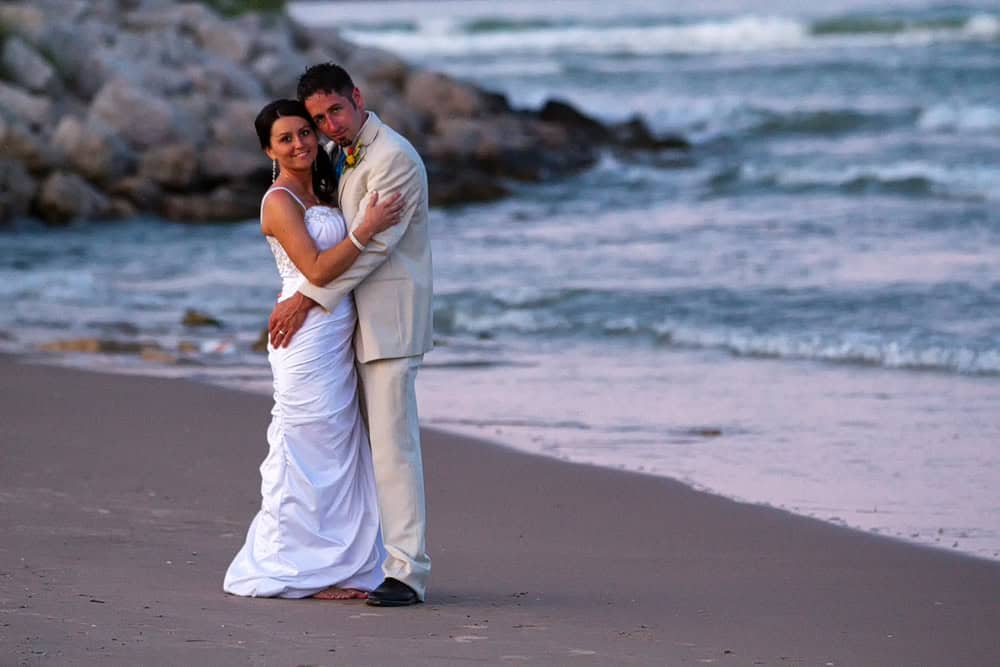 A bride and groom standing on the beach at sunset.