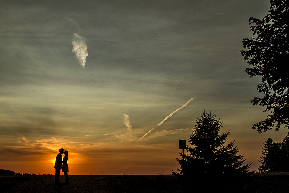 A country engagement photo of a couple standing in a field at sunset.