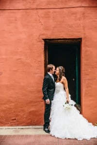 A bride and groom kiss in front of a red building.