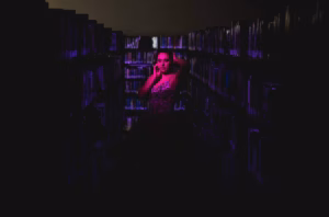 A person stands between bookshelves in a dimly lit library, illuminated by purple light, holding a phone to their ear—captured with the creative touch of a La Crosse Senior Photographer.