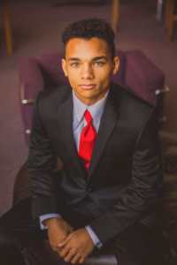 A young man in a black suit with a red tie sits on a dark chair, looking directly at the camera with a neutral expression, captured by a talented La Crosse Senior Photographer.
