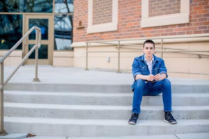 A young person in a denim jacket and jeans sits alone on concrete steps outside a brick building with metal railings—captured by a talented La Crosse Senior Photographer.