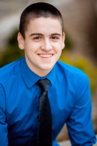 A young man with short dark hair, wearing a bright blue dress shirt and black tie, smiles at the camera outdoors—captured by a La Crosse Senior Photographer.
