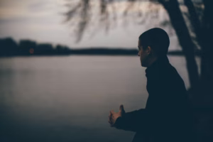A person with short hair stands by a body of water at dusk, looking out over the calm surface with trees in the background—captured beautifully by a La Crosse Senior Photographer.