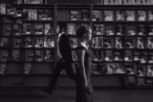 A woman in a dress stands in the foreground while a man in a suit walks past her by magazine shelves at a library or bookstore, captured in the candid style of a La Crosse Senior Photographer.