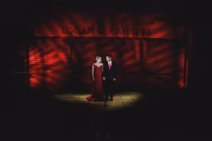 A woman and a man in formal attire stand holding hands under a spotlight in a gymnasium with "LOGAN RANGERS" on the wall and red lighting effects, captured by a La Crosse Senior Photographer.
