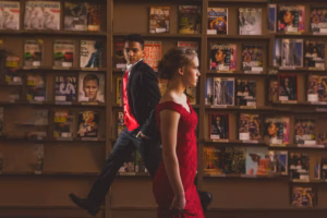 A young woman in a red dress walks past a man in a suit with a red vest in front of a wall of magazines, captured with the keen eye of a La Crosse Senior Photographer.