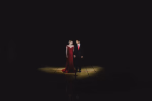 A woman in a red dress and a man in a black suit stand holding hands under a spotlight on a dark stage, captured beautifully by La Crosse Senior Photographer.