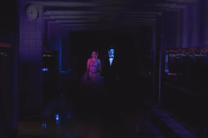 A man in a suit and a woman in a formal dress stand under dramatic blue and pink lighting in a dark hallway with a clock showing 4:07, captured by a talented La Crosse Senior Photographer.