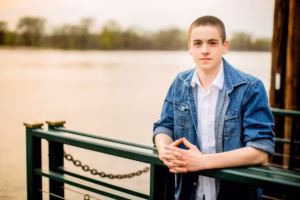 A young person with short hair, wearing a denim jacket and white shirt, leans on a green metal railing by a body of water with trees in the background, captured beautifully by a La Crosse Senior Photographer.