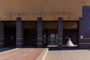 A bride and groom stand together outside the entrance of Holmen High School on a sunny day, captured beautifully by a La Crosse Senior Photographer.