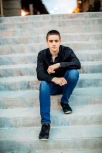 Young man in a black shirt and blue jeans sits on outdoor concrete steps, looking at the camera with a neutral expression—captured by a La Crosse Senior Photographer.