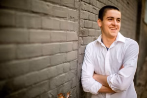 Young man in a white shirt stands with arms crossed, smiling, next to a gray brick wall—captured by a La Crosse Senior Photographer.