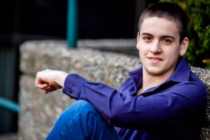 A young man with short dark hair, wearing a dark purple shirt and blue jeans, sits outdoors against a stone wall, looking at the camera and smiling slightly—captured perfectly by a La Crosse Senior Photographer.
