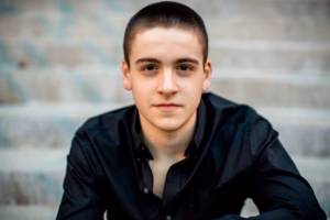 A young person with short dark hair, wearing a black button-up shirt, sits on outdoor steps and looks directly at the camera—captured by a talented La Crosse Senior Photographer.