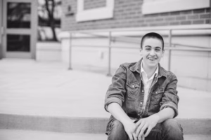 A young person with short hair, wearing a denim jacket, sits on outdoor steps and smiles. Captured in black and white by a La Crosse Senior Photographer.