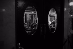 Black and white photo of people seated in a large room, viewed through oval windows in double doors, suggesting a formal or governmental setting—perhaps even a Wisconsin Capital Wedding.