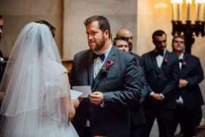 A groom in a tuxedo reads vows to his bride in a wedding dress and veil during a touching Wisconsin Capital Wedding ceremony, with groomsmen standing in the background.