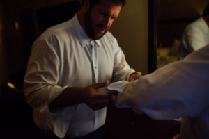 A man in a white dress shirt helps another person adjust their sleeve in a dimly lit room, preparing for a Wisconsin Capital Wedding.