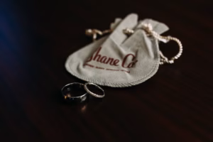 Two rings, one plain and one adorned with small stones, rest on a dark surface beside a light-colored jewelry pouch labeled "Shane Co."—a timeless symbol perfect for a Wisconsin Capital Wedding.