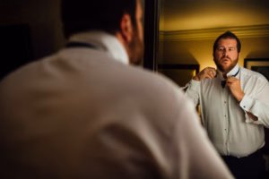 A man in a white dress shirt stands in front of a mirror, adjusting his bow tie, preparing for his Wisconsin Capital Wedding, with his reflection visible.
