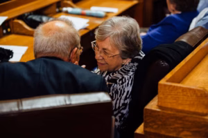 An elderly man and woman sit closely together in wooden seats, engaged in conversation, inside what appears to be a formal setting at the Wisconsin Capital, perhaps attending a wedding or legislative event.