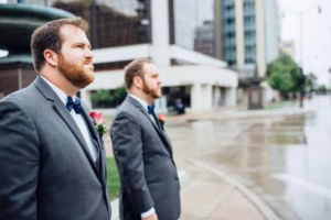 Two men in suits with boutonnieres stand on a wet sidewalk near modern buildings, gazing into the distance after their Wisconsin Capital Wedding.
