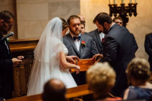 A bride and groom, assisted by a man, lock a wooden box during their Wisconsin Capital Wedding ceremony inside a church, with guests seated in the foreground.