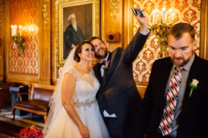 A bride and groom in wedding attire take a selfie together indoors at their Wisconsin Capital Wedding, while a man in a suit walks by in front of them.