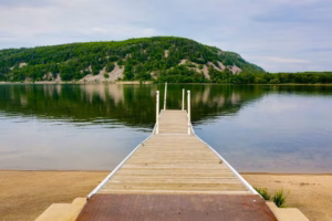 A wooden dock extends over the calm waters of Devils Lake, with a sandy shore in the foreground and a tree-covered hill rising under a partly cloudy sky.