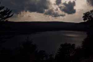 A dark, cloudy sky looms over the calm waters of Devils Lake, surrounded by silhouetted trees and distant hills.