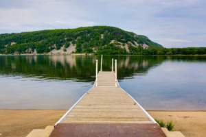 A wooden dock extends over the calm waters of Devils Lake toward a green, tree-covered hill under a cloudy sky. Sand surrounds the dock at the shoreline.