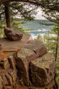 Large, flat rock outcrop with pine trees overlooking a forested area and Devils Lake shimmering in the background.