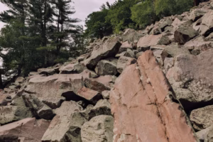 Large, rugged rocks and boulders are scattered across a hillside at Devils Lake, with trees and greenery in the background beneath a cloudy sky.