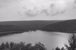 A calm lake, known as Devils Lake, is surrounded by tree-covered hills under a cloudy sky, viewed from an elevated vantage point.