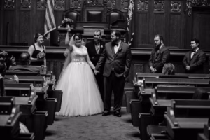 A bride and groom stand at the front of a Wisconsin Capital Wedding courtroom, holding hands, while guests and wedding party members look on. The bride raises her bouquet in celebration.