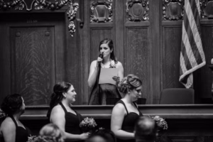 A woman stands at a podium reading from a paper into a microphone during a Wisconsin Capital Wedding, while three women in matching dresses holding bouquets stand in the foreground.