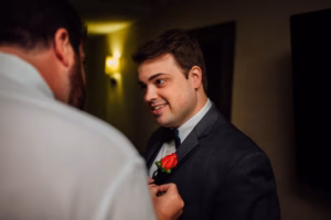 A man in a suit smiles as another pins a red rose boutonniere to his lapel indoors under warm lighting, capturing a joyful moment at a Wisconsin Capital Wedding.