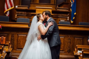 Bride and groom share a kiss at the altar in a courthouse setting, surrounded by wooden benches, with flags in the background—a charming Wisconsin Capital Wedding moment.