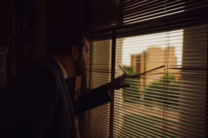 A man in a suit stands indoors, peering outside through the window blinds at the daylight and buildings—perhaps anticipating a special moment like a Wisconsin Capital Wedding.