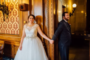 A bride and groom stand on opposite sides of a wooden wall at their Wisconsin Capital Wedding, holding hands around the corner without seeing each other.