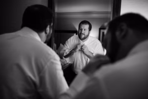 A man is buttoning his shirt while looking at himself in a mirror, preparing for a Wisconsin Capital Wedding; his reflection is clearly visible.