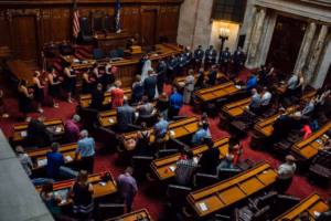 People are seated and standing in a large, formal legislative chamber with wood paneling, desks, and flags, suggesting a government or official event—such as a Wisconsin Capital Wedding—is taking place.
