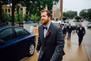 A man in a suit with a red rose boutonniere walks on a wet sidewalk near parked cars, as other formally dressed people gather near city buildings—a classic moment from a Wisconsin Capital Wedding.
