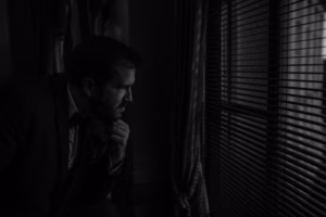 A man in a suit looks thoughtfully out a window with closed blinds, resting his chin on his hand in a dimly lit room, perhaps reflecting on an unforgettable Wisconsin Capital Wedding.