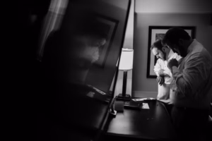 Two men in dress shirts stand by a desk in a dimly lit room, adjusting their attire for a Wisconsin Capital Wedding, framed pictures and a lamp casting soft light around them.