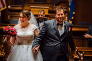 A bride and groom holding hands and smiling while walking down the aisle in a Wisconsin Capital Wedding courtroom setting.