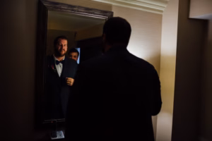 A man in a suit stands in front of a mirror, adjusting his jacket, with another person partially visible in the reflection behind him at a Wisconsin Capital Wedding.