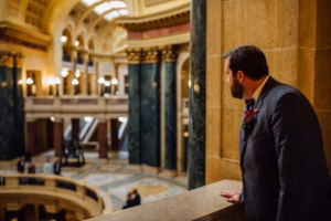 A man in a suit stands at a marble balcony inside the ornate Wisconsin Capital, with columns, arches, and decorative lighting—a stunning setting for a wedding as he gazes toward the grand interior.