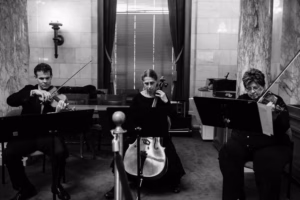 Three musicians perform indoors at a Wisconsin Capital Wedding; two play violins and one plays a cello, all seated in formal attire with sheet music stands in front of them.